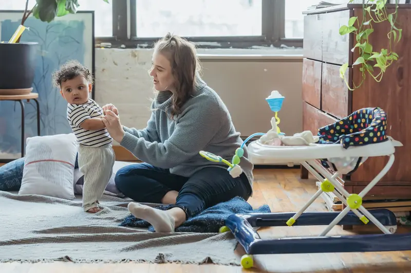 Full length positive young mother in casual clothes and adorable ethnic baby holding hands and playing together on floor in light living room