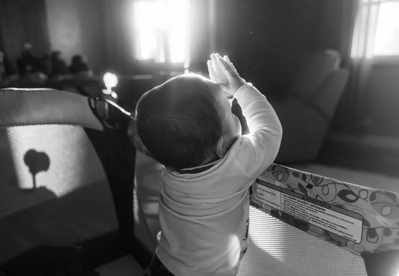 A baby stands in a crib with hands in prayer in a sunlit room, captured in black and white