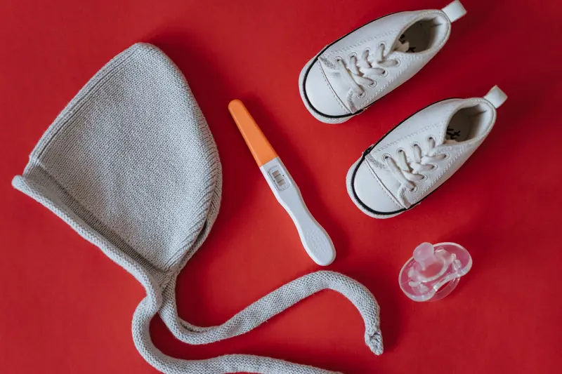 A flat lay image featuring baby shoes, a pregnancy test, and baby essentials on a red background