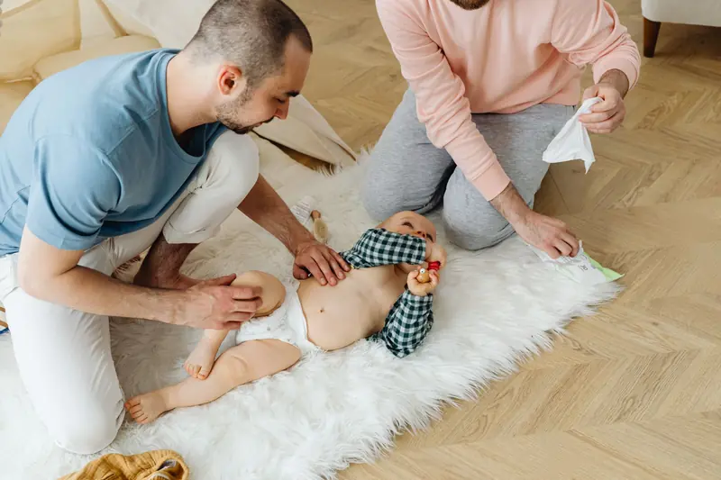 Two fathers changing their baby's diaper on a soft white rug at home, demonstrating parenting and togetherness