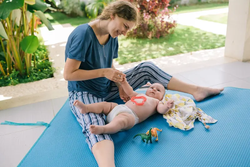 Smiling mother and baby play on a mat outside, enjoying a sunny day with toys