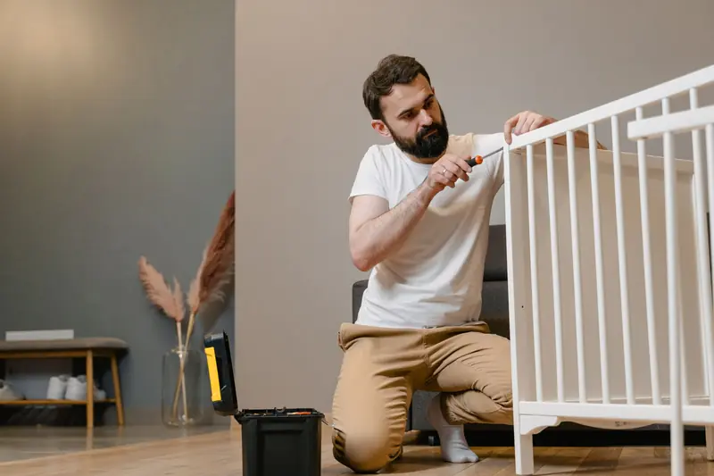 Father kneels to fix a white baby crib using a screwdriver in a contemporary nursery setting